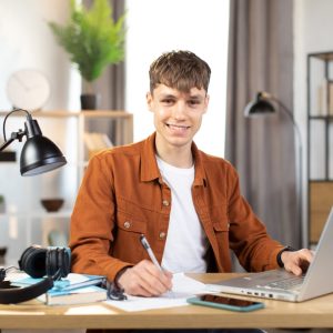 Happy,Caucasian,Student,In,Casual,Wear,Sitting,At,Table,With student z laptopem przy biurku